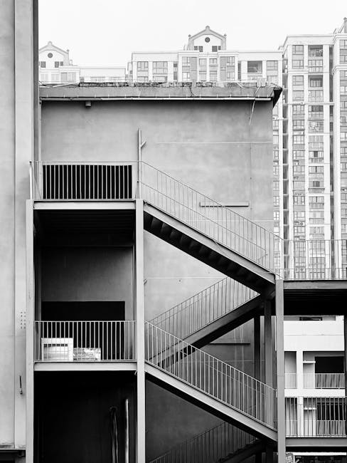 A black-and-white photograph of an external metal fire escape staircase attached to the side of a multi-storey building. The staircase features three flights of stairs with metal railings, connecting